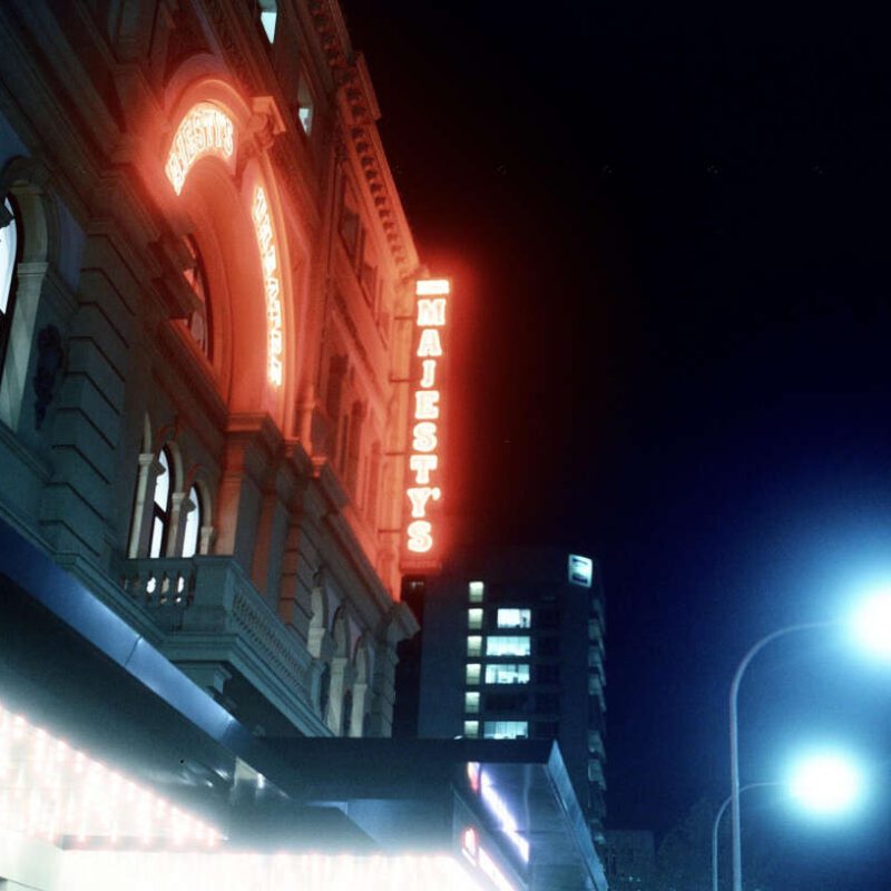 Exterior of a theatre building at night with red neon signs reading "Majesty's," and illuminated windows and streetlights.