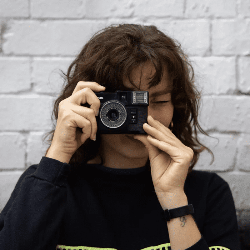 Person holding a camera in front of their face, standing against a brick wall.
