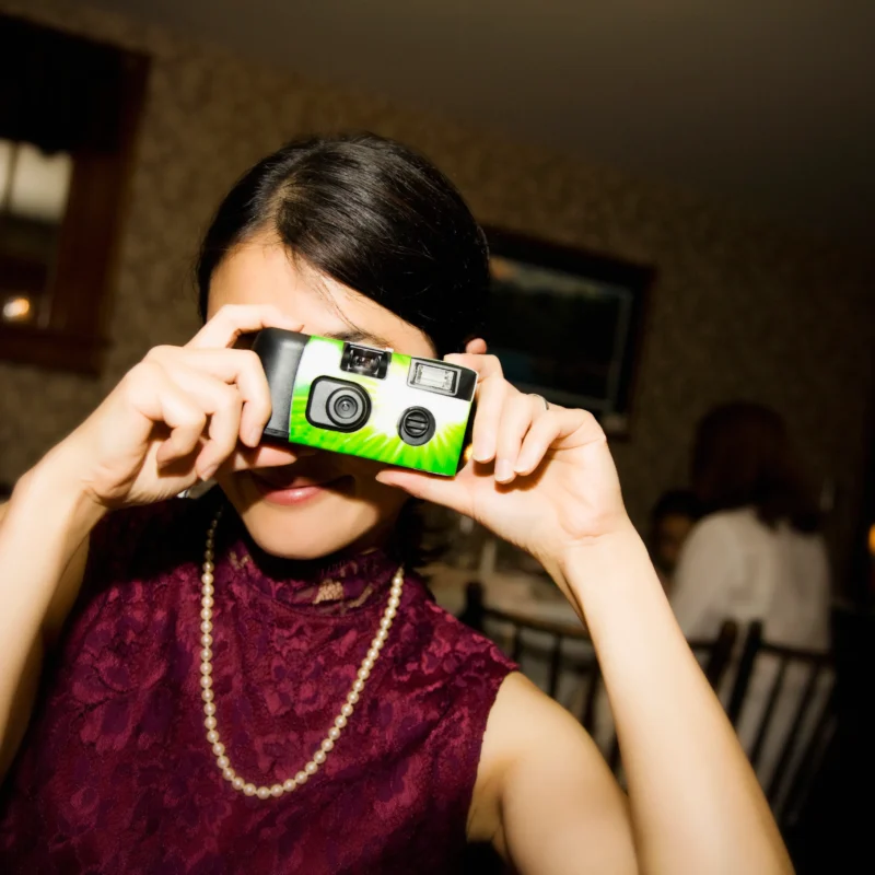 A woman holding a disposable camera to her face, wearing a purple lace dress and pearl necklace, in a dimly lit room.