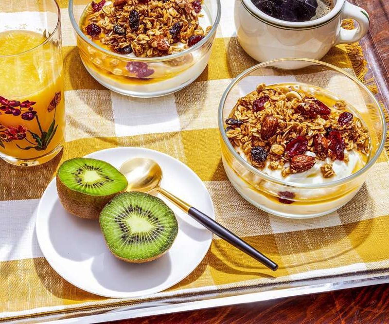 Breakfast setup with granola and yoghurt bowls, orange juice, coffee, and kiwi slices on a checkered tablecloth.