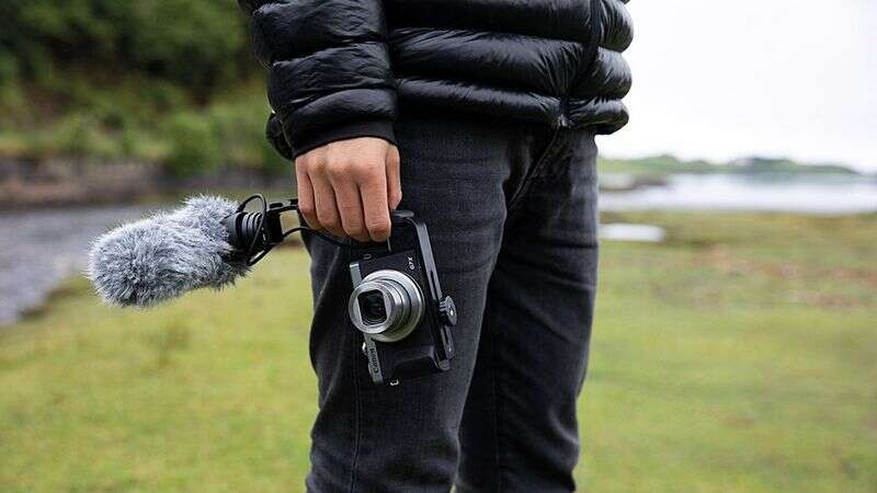 Person holding a camera with a mounted external microphone, standing outdoors on a grassy area.