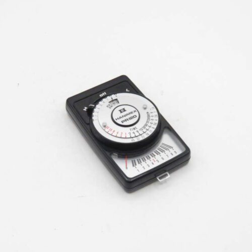 Black analog light meter with a dial and digital readout, branded "Hanımex RİXO PR120," lying on a white surface.
