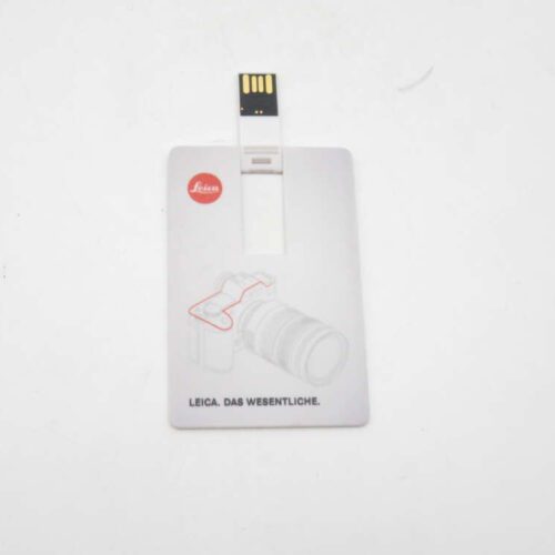 A white USB flash drive in the shape of a card with a Leica logo and the text "LEICA. DAS WESENTLICHE." along with a faint outline of a camera graphic.