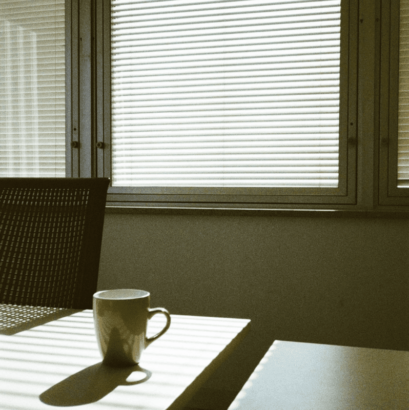 A white coffee cup on a table next to a chair, with sunlight streaming through window blinds, casting striped shadows.