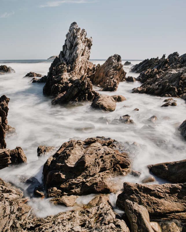 Rocks on a shoreline with waves crashing against them under a clear sky.