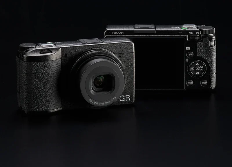 Two black Ricoh compact cameras, one showing the lens and the other showing the rear controls and screen, against a dark background.