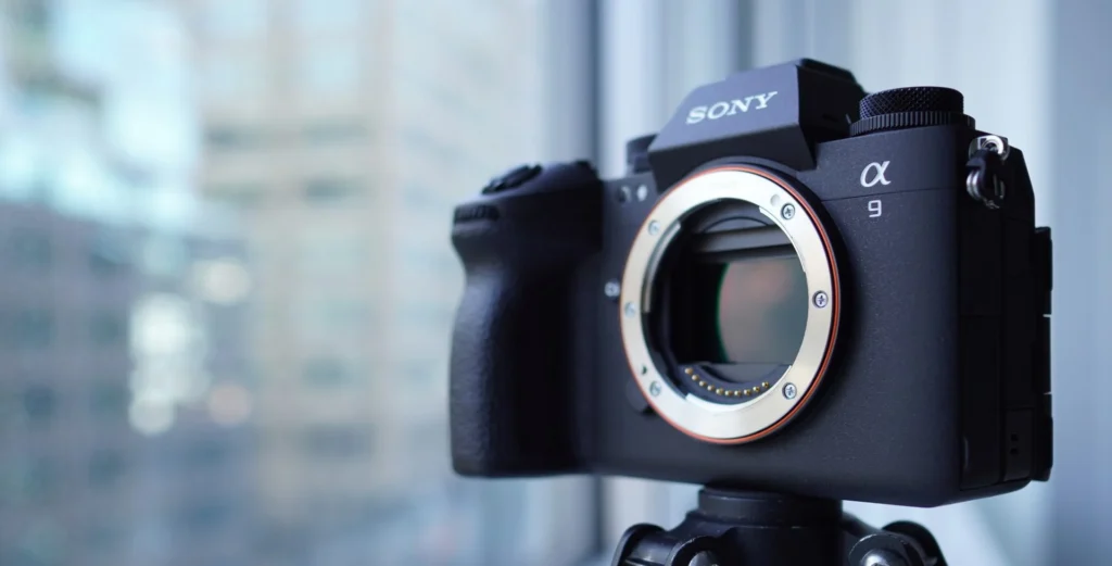 A Sony Alpha 9 camera on a tripod, without a lens, with blurred city buildings in the background.