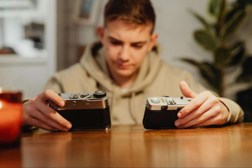 Young person in a hoodie holding two vintage cameras, sitting at a wooden table.