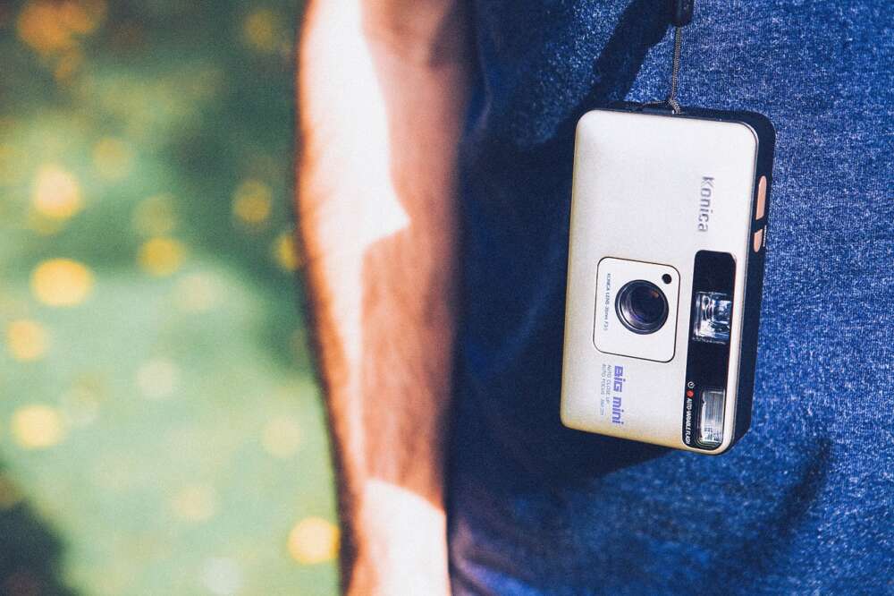 A small, retro Konica Big Mini camera hangs on a strap around a person's neck, resting against a blue shirt.