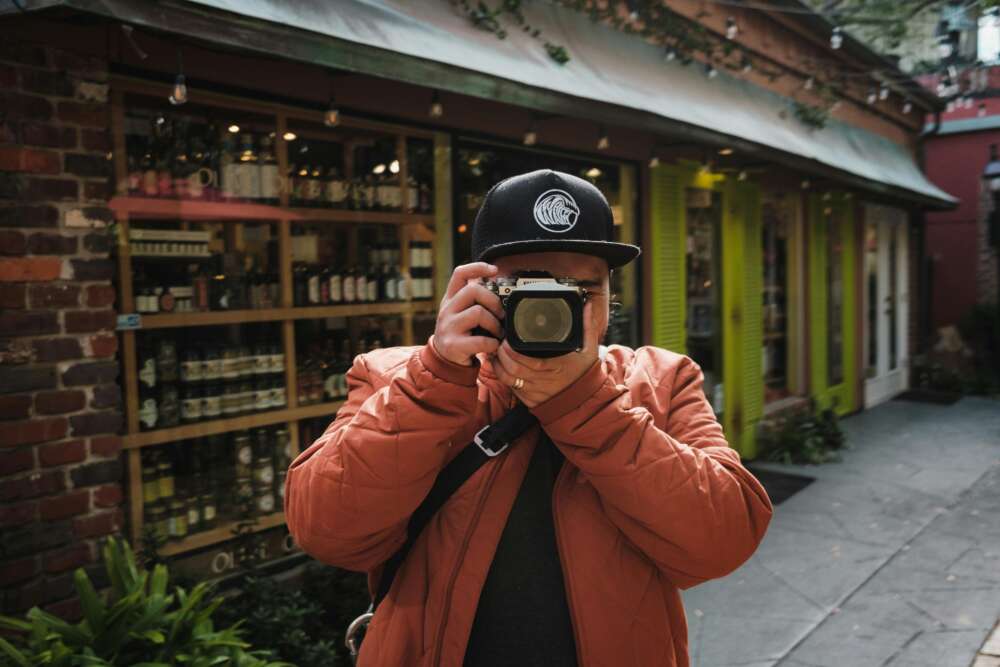 Person in an orange jacket and cap taking a photo on a city street.