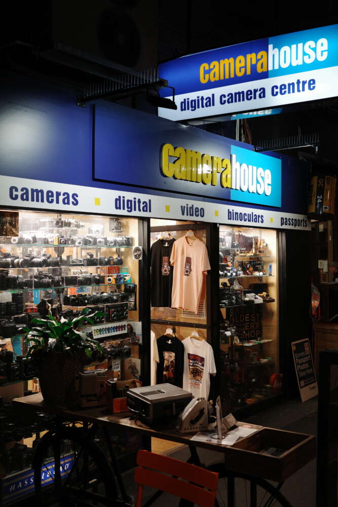 Camera store front displaying cameras, video equipment, and branded shirts under signage that reads "camera house digital camera centre."