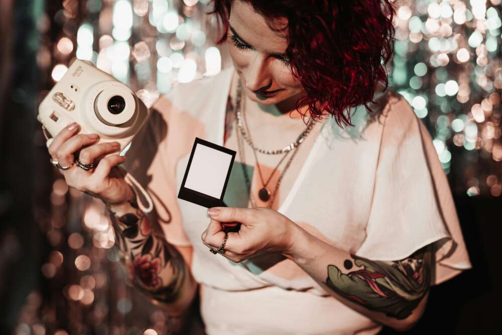 A woman with red curly hair and arm tattoos holding an instant camera and a blank photo, against a sparkling background.