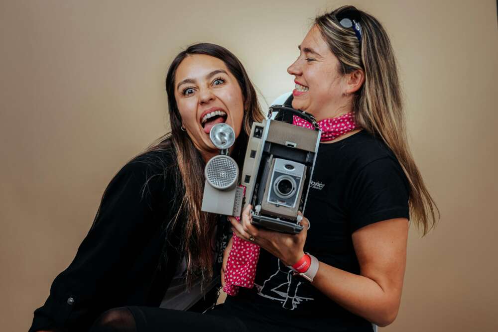 Two women laughing and playfully posing with an old camera against a plain background.