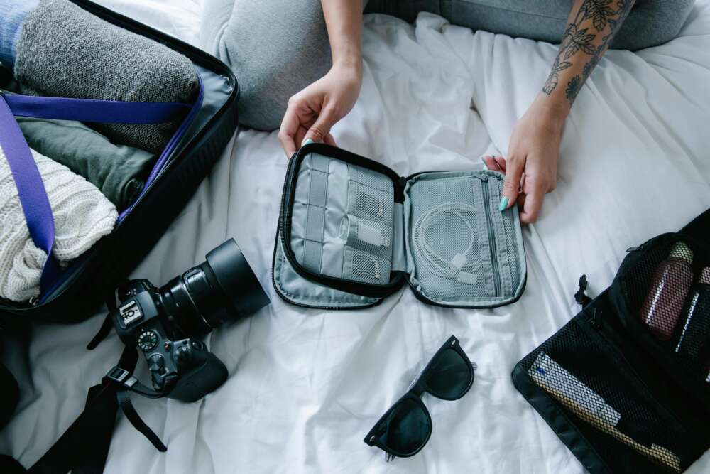 Person packing a suitcase with clothes, a camera, a pair of sunglasses, and an open organiser with cables on a bed.