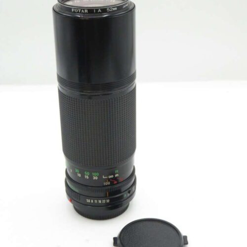 A black Fotar 100-200mm 1:4.5 camera lens with a 52mm filter size, standing upright next to a detached lens cap.