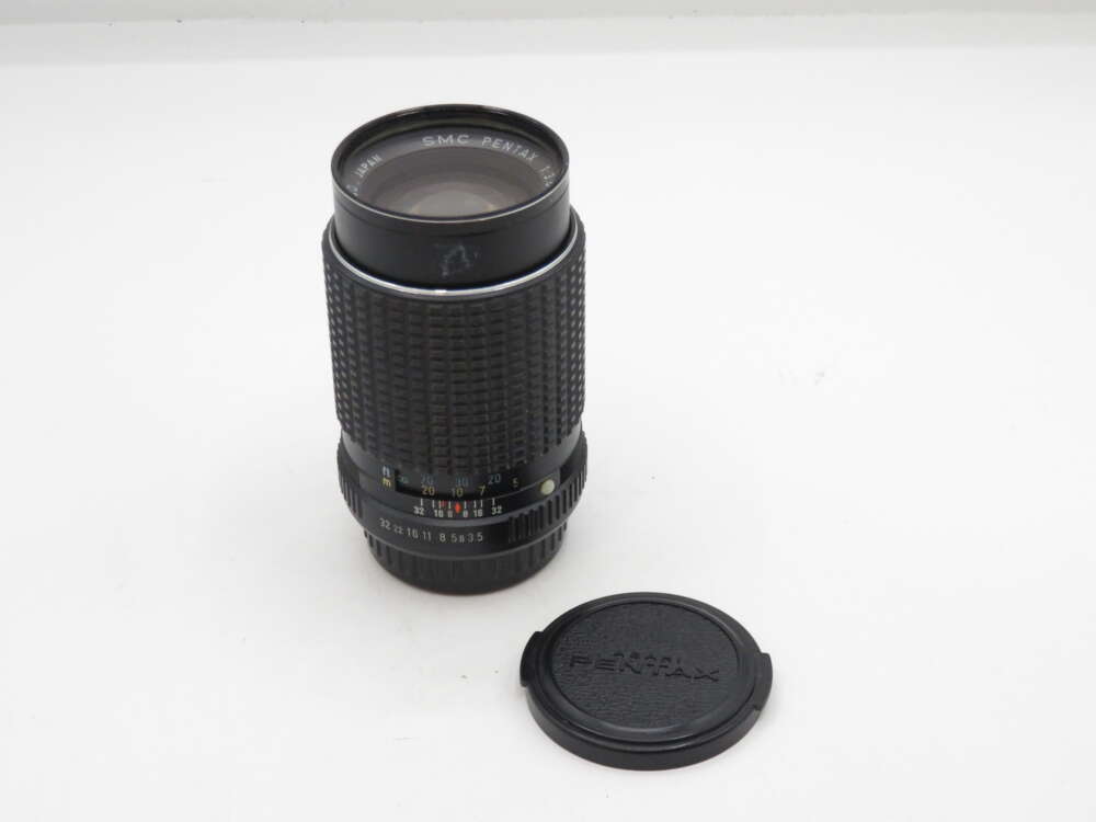 IMG_0354 A black SMC Pentax camera lens with its detached lens cap placed beside it on a white surface.