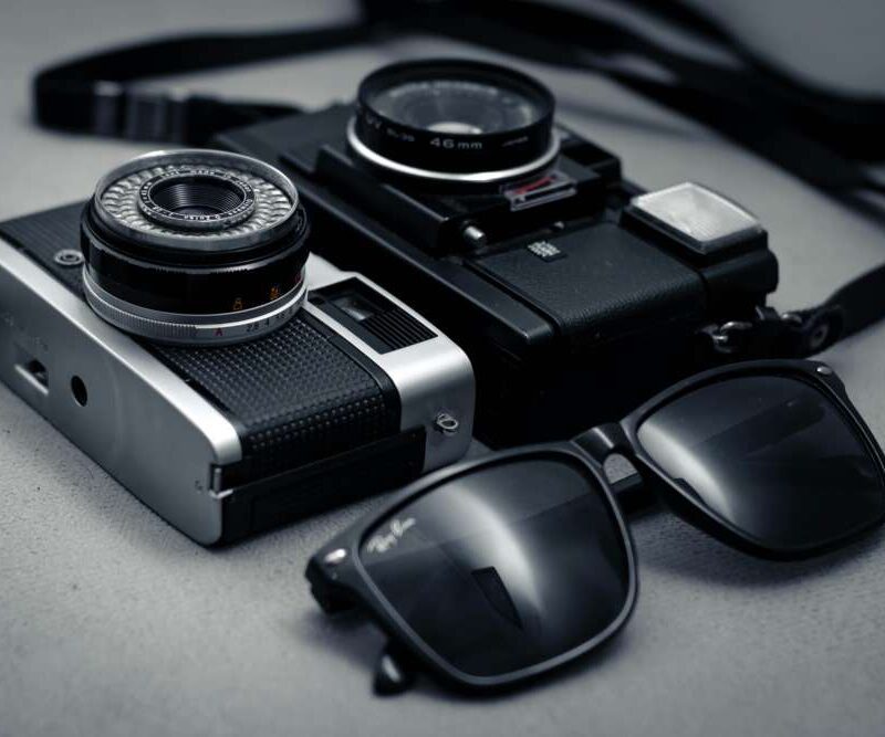 Two vintage film cameras alongside a pair of black sunglasses on a grey surface.
