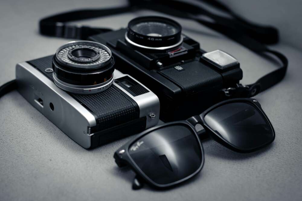 Two vintage film cameras alongside a pair of black sunglasses on a grey surface.