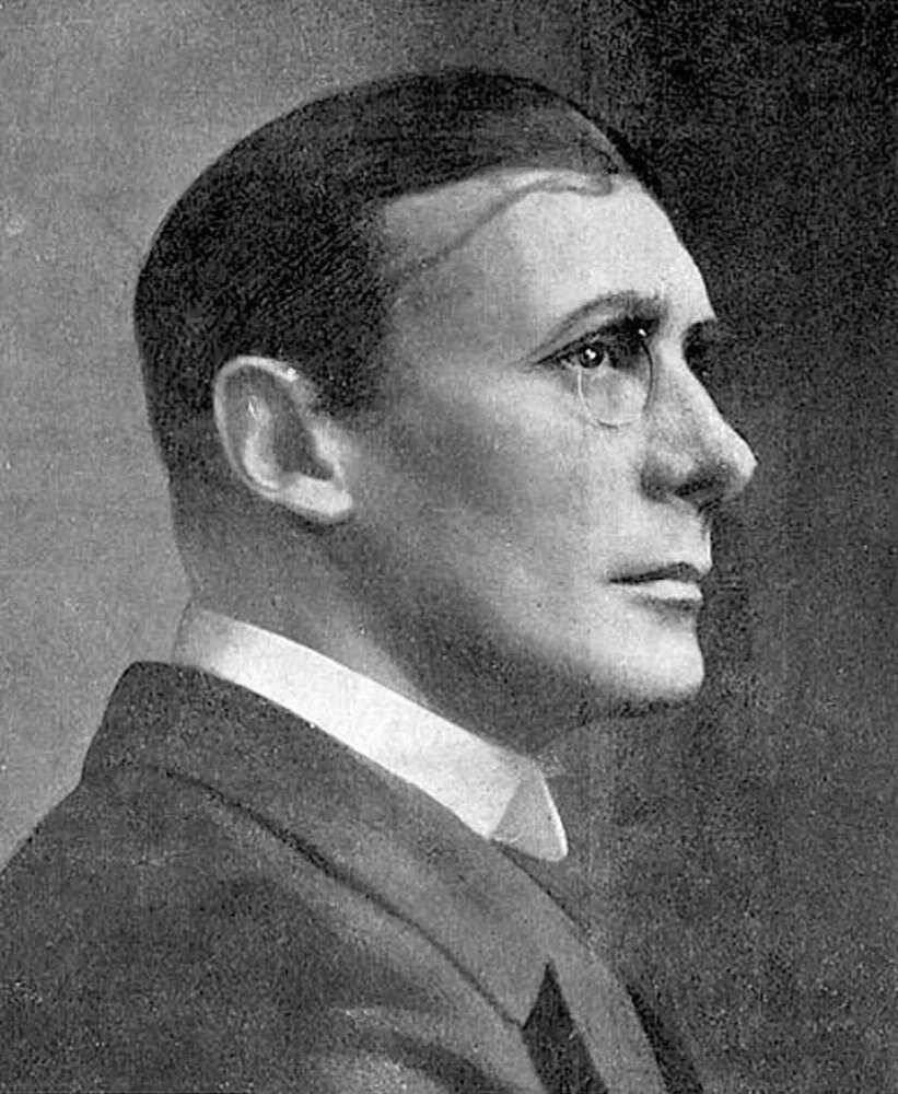 Portrait of John Wade in profile wearing a suit and glasses, with slicked-back hair.