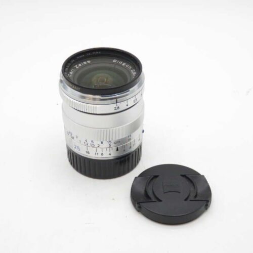 A Carl Zeiss Biogon 28mm camera lens with a silver and black body, placed next to a detached black lens cap on a white surface.