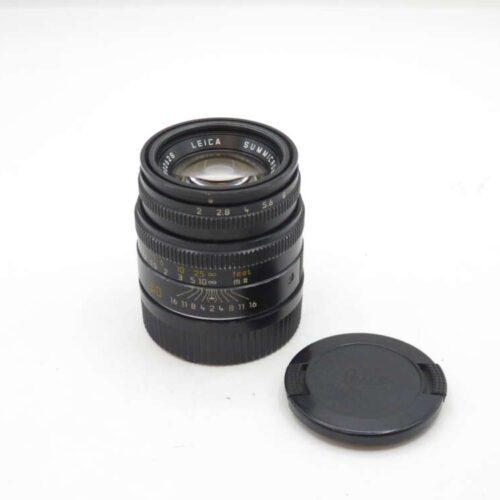 A black Leica camera lens with an attached zoom and focus ring, placed upright next to a detached lens cap on a white surface.