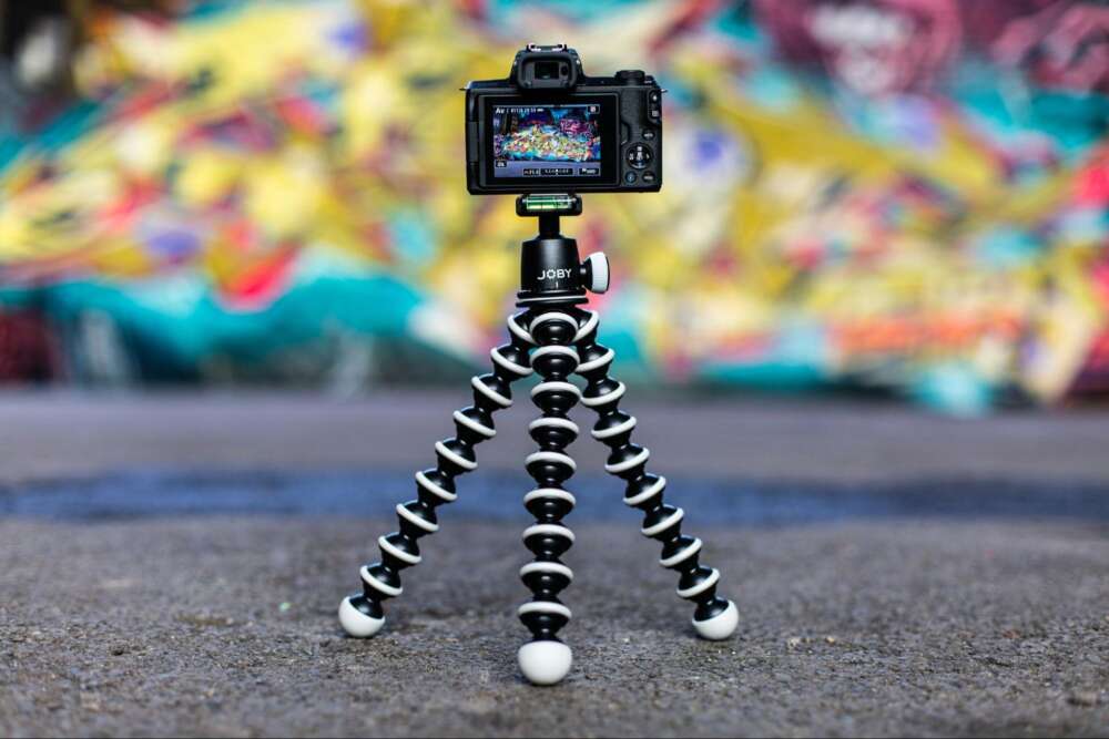 A digital camera mounted on a flexible Joby tripod, positioned on the ground with a colourful, blurred graffiti wall in the background.