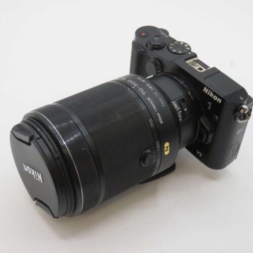 A Nikon 1 V3 camera with a large zoom lens attached, placed on a white surface.