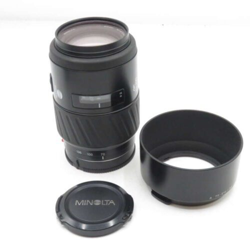 A Minolta camera lens with a separate lens hood and cap, featuring a zoom range of 70-210mm, placed on a white surface.