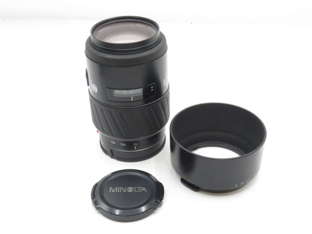 IMG_0281 A Minolta camera lens with a separate lens hood and cap, featuring a zoom range of 70-210mm, placed on a white surface.