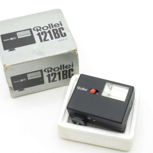 A Rollei 121 BC camera flash unit with its original packaging. The flash is black with a red button and a clear lens, sitting in a white foam base. The packaging, in the background, is gray with product branding.