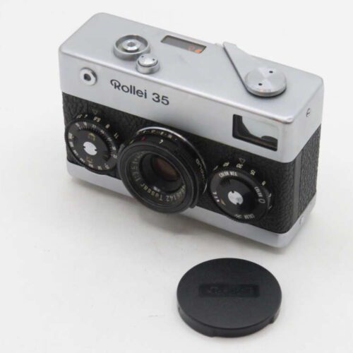 A Rollei 35 camera with a silver and black body, featuring a Tessar 3.5/40 lens and control dials, resting on a white surface with a detached lens cap placed in front.
