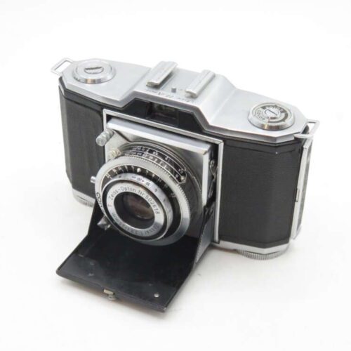 A vintage foldable camera with a black and silver body, featuring a collapsible bellows and a prominent lens in the centre. The camera is positioned on a white background.