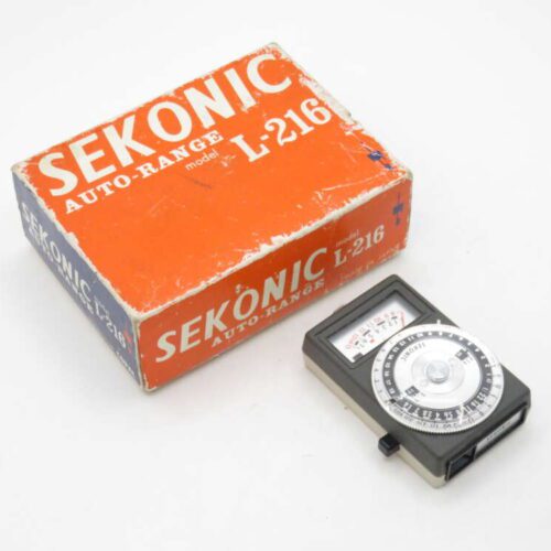 A Sekonic Auto-Range light meter model L-216 with its original orange and white box. The light meter features a circular dial on a rectangular body, positioned beside the box on a white background.