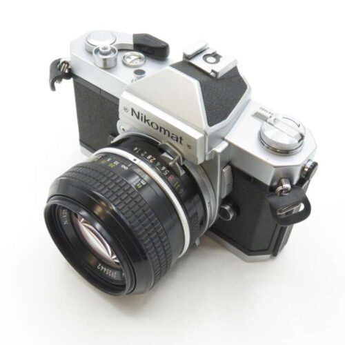 A silver and black Nikomat film camera with a detachable lens, featuring classic SLR design elements and a viewfinder on top.