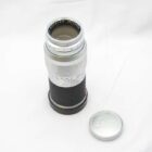 A vintage Leica camera lens with a silver and black body, featuring aperture and focus markings. The lens cap is placed next to it on a white surface.