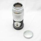 A vintage Leica Elmarit camera lens with a silver finish and black focus ring, sitting on a white surface next to a silver Leica lens cap.