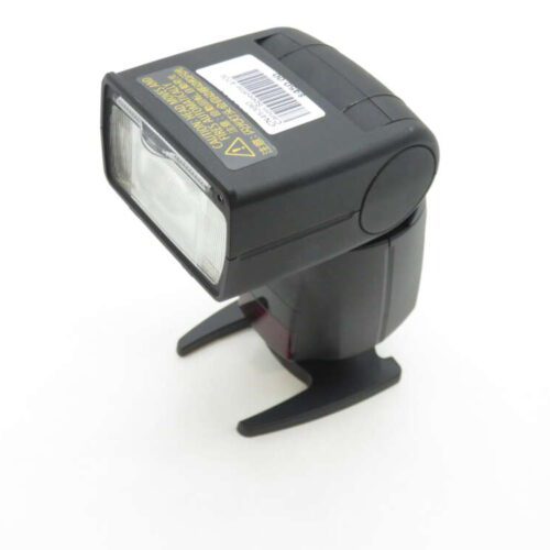 A black external camera flash with a rectangular flash head and a stand, placed on a white surface.