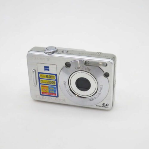 A silver Sony Cyber-shot digital camera featuring a Carl Zeiss lens, 6.0 megapixels, and product stickers detailing features like effective ISO sensitivity and battery life.