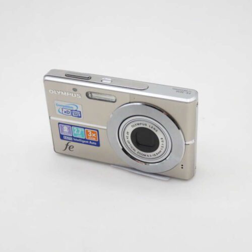 A silver Olympus digital camera with a compact design, featuring a 3x optical zoom lens and various stickers indicating features such as Intelligent Auto and compatibility with xD and microSD cards.