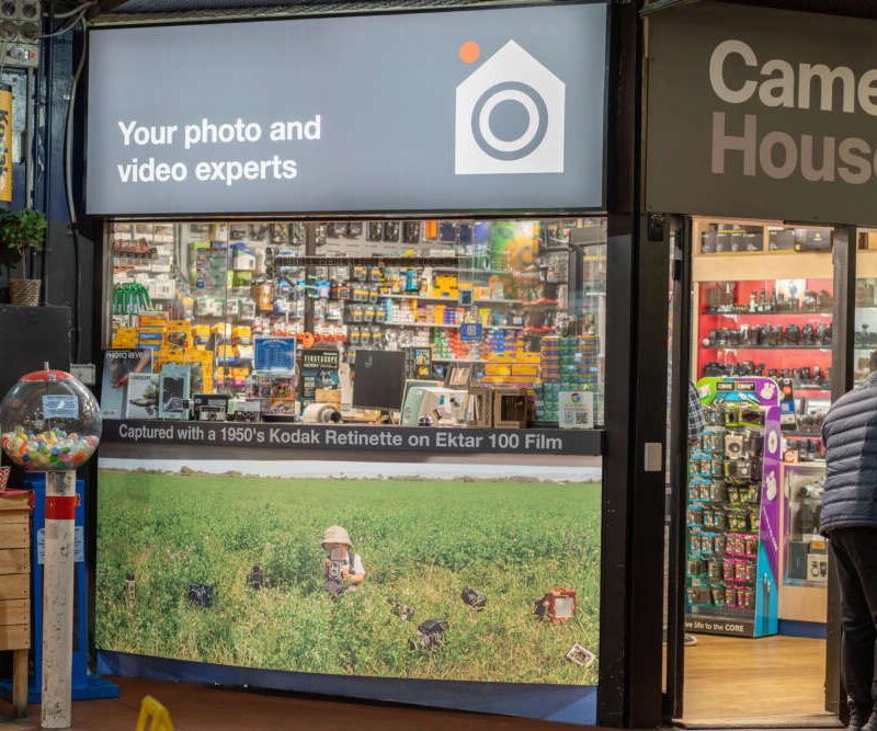 A storefront of a camera store named 'Camera House' with a sign that reads 'Your photo and video experts.' The window display features various camera equipment and a large photograph of a person in a field capturing images. A man in a jacket is examining products inside the shop.
