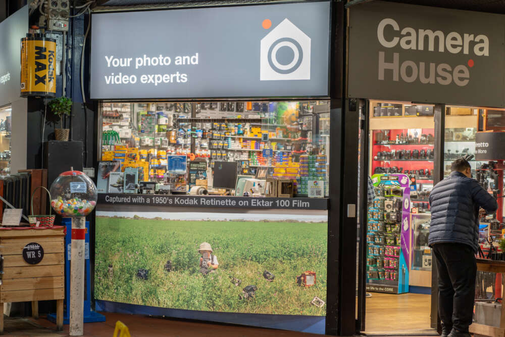 A storefront of a camera store named 'Camera House' with a sign that reads 'Your photo and video experts.' The window display features various camera equipment and a large photograph of a person in a field capturing images. A man in a jacket is examining products inside the shop.