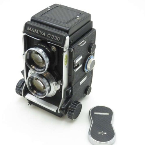 A Mamiya C330 twin-lens reflex camera positioned on a light background, showing its dual lenses and the black textured body with various control dials. Beside the camera, there is a black and silver lens cap resting on the surface.
