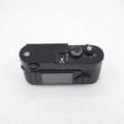 A black Leica camera viewed from above, showing its top and back panels with control dials, a small screen, and a circular dial with shutter speed settings. The camera's textured grip and compact design are visible.