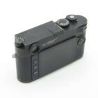 A black camera with a textured grip, featuring a rear LCD screen and various control dials on top, labeled "LEICA CAMERA WETZLAR MADE IN GERMANY."