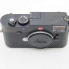 A Leica camera with a black body, featuring manual dials and a lens cap, viewed from the top at an angle.