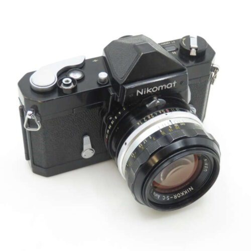 A black Nikomat vintage film camera with a prominent lens, featuring various control dials and a textured grip, placed against a white background.