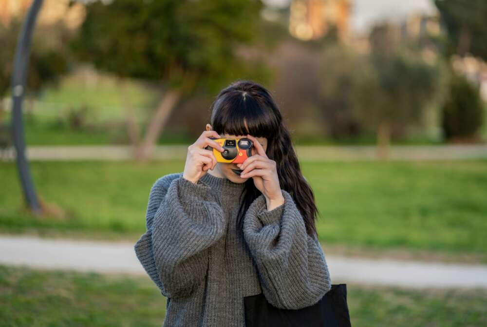 A person in a grey sweater holds a small yellow and black camera to their face in a park setting with greenery in the background.