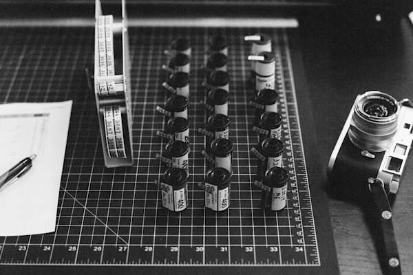 A black and white image showing a grid cutting mat with rows of camera film rolls, a vintage camera, a pen, and a piece of paper, creating a workspace for film photography.