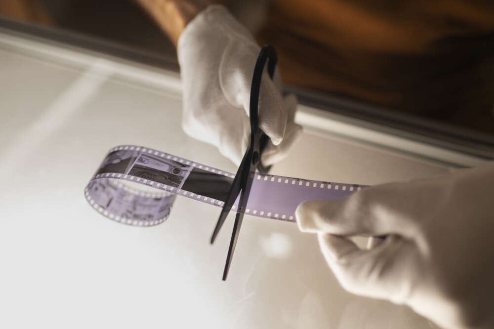 Hands wearing white gloves cutting a strip of photographic film on a lightbox with scissors, highlighting the film's perforated edges and negative images.