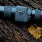 A Kite Optics telescope lying on a tree trunk with a yellow leaf nearby.
